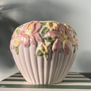 May include: A white ceramic planter with a scalloped rim and a floral design. The planter features pink and yellow flowers with green leaves.