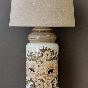 May include: A table lamp featuring a cylindrical white ceramic base decorated with hand-painted beige and brown floral motifs. The lamp is topped with a textured, light beige fabric drum lampshade and includes a white power cord at the base.