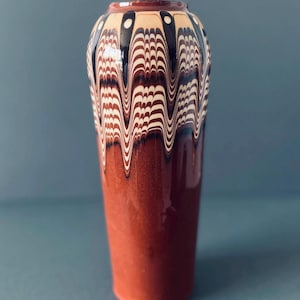 Könnte beinhalten: Eine zylindrische Keramikvase mit glatter, glänzender Oberfläche. Der untere Teil ist in einem einfarbigen, tiefen Rotbraun gehalten, während der obere Bereich ein dekoratives Marmormuster aus cremefarbenen, schwarzen und braunen Wellenlinien zeigt.
