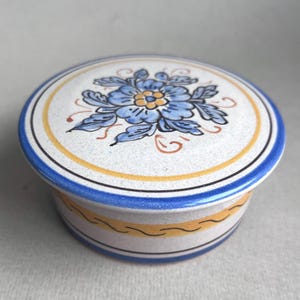 Può includere: Una scatola rotonda in ceramica per bigiotteria con un design floreale blu e giallo. Il coperchio ha un fiore blu con un centro giallo e foglie gialle. La scatola ha una fascia blu e gialla attorno alla parte inferiore.