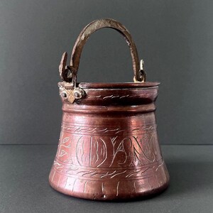 May include: A small, tapered copper bucket featuring a weathered metal swing handle attached with rivets. The exterior surface displays a series of horizontal bands and intricate, hand-etched decorative patterns encircling the body.