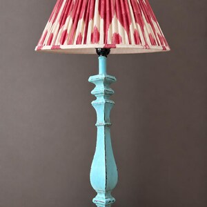 May include: A table lamp featuring a light blue painted wooden base with a distressed finish and a pleated lampshade with a red and cream ikat-style diamond pattern. The lamp has a square base and a black power cord extending from the side.