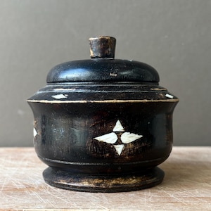 May include: A small, round, dark brown wooden lidded container featuring a smooth, polished finish. The exterior is decorated with inlaid white, diamond-shaped accents arranged in a star pattern around the centre and along the upper rim.