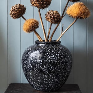 Puede incluir: Un jarrón de cerámica negro y redondo con un patrón de motas blancas que contiene cinco cabezas de flores secas de color marrón en forma de globo sobre tallos leñosos finos. El jarrón tiene un acabado liso y brillante, un cuerpo bulboso y una abertura.