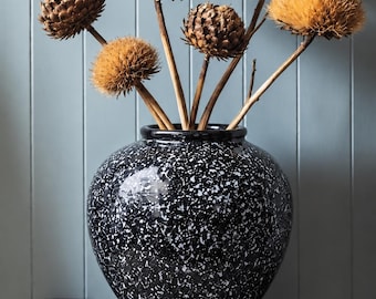 May include: A round, black ceramic vase with a white speckled pattern containing five dried, brown, globe-shaped flower heads on thin, woody stems. The vase has a smooth, glossy finish and a wide, bulbous body with a narrow, circular opening at the top.