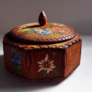 Puede incluir: Una caja redonda de madera con una tapa que presenta un diseño floral pintado a mano, incluyendo una flor de edelweiss blanca y flores azules. La caja tiene un borde festoneado tallado en la parte superior y un pequeño pomo de madera.