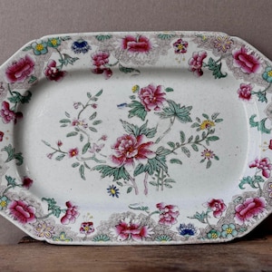 May include: An oval ceramic serving platter featuring a cream-colored base with a central floral motif of pink peonies and green leaves. The wide border is decorated with a repeating pattern of pink flowers, small blue blossoms, and intricate brown lace-like detailing.