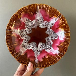 Peut inclure: Une assiette décorative en céramique avec un motif en forme d'éclat d'étoile. L'assiette présente un bord marron avec un bord festonné, entourant un centre de glaçure marron, rose et blanche. L'assiette est tenue par une main sur un fond gris.
