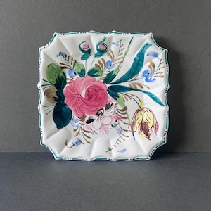 Puede incluir: Plato cuadrado de cerámica blanca con borde festoneado, pintado a mano con un diseño floral. Las flores son principalmente rosas, con detalles verdes, azules y amarillos. El borde está delineado en verde azulado.