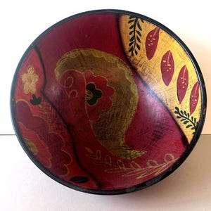 Puede incluir: Un cuenco decorativo de madera con interior rojo oscuro y borde negro. El cuenco presenta diseños florales y de hojas dorados y rojos. Los diseños dorados tienen un aspecto desgastado y texturizado.