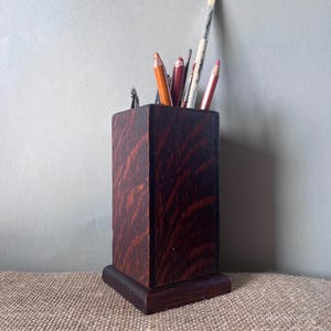 Pode incluir: Um porta-lápis de madeira escura com formato retangular e base. O suporte contém vários materiais de arte, incluindo lápis e pincéis. O veio da madeira é visível e a cor é marrom avermelhado escuro.
