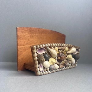 Puede incluir: Un organizador de escritorio de madera con un panel frontal rectangular decorado con varias conchas marinas. Las conchas son en tonos de blanco, marrón y morado, y están delineadas con pequeñas cuentas redondas de color claro.