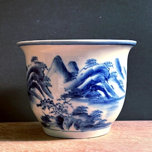 May include: A white ceramic planter featuring a hand-painted blue landscape design. The exterior displays detailed illustrations of mountains, trees, and water, with a smooth, flared rim and a tapered base, creating a classic vessel shape.