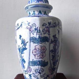Puede incluir: Un tarro de jengibre de cerámica blanca con tapa extraíble, decorado con motivos florales y de aves pintados a mano en azul y rosa. El recipiente tiene un cuello estrecho y un cuerpo ancho decorado con paneles verticales con ilustraciones.