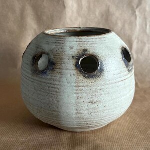 May include: A spherical ceramic vase with a textured, off-white matte finish. The vessel features multiple circular openings around its upper circumference, each accented with dark, charred-looking edges that contrast with the light body.