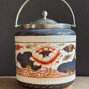 Può includere: Un barattolo cilindrico in ceramica per biscotti con coperchio e manico basculante in metallo argentato. Il corpo presenta uno sfondo bianco con fiori di loto arancioni, foglie blu scuro stilizzate e dettagli dorati, bordato da motivi a greca in rilievo.