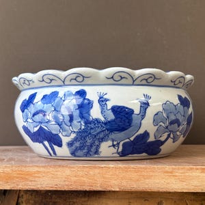 Peut inclure: Un bol en céramique rond présentant une base blanche avec des illustrations peintes à la main en bleu d'un paon et de grandes fleurs. Le bol possède un bord festonné décoré d'un motif de ligne bleue continue sur le pourtour supérieur.