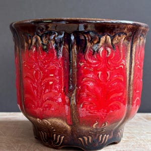 Puede incluir: Una maceta de cerámica con un esmalte rojo vibrante y detalles en marrón oscuro tipo goteo en el borde y la base. La superficie exterior está decorada con un patrón floral en relieve repetitivo y crestas verticales texturizadas.