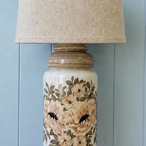May include: A table lamp with a cylindrical ceramic base featuring a hand-painted floral pattern in shades of brown, tan, and cream. The lamp is topped with a textured, light beige fabric drum shade and has a smooth, glossy finish.