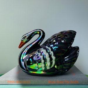 May include: A black iridescent ceramic swan figurine with white accents. The swan has a red beak and is sitting on a stack of books. The books have the text "Music From The Studio" and "ONEY-SUCKLE COMPANY spiritus" on the covers.