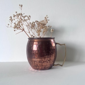 May include: A copper Moscow Mule mug with a hammered finish and a brass handle. The mug is filled with dried flowers. The copper has a reddish-brown hue, and the handle is a gold colour. The mug is sitting on a white surface.