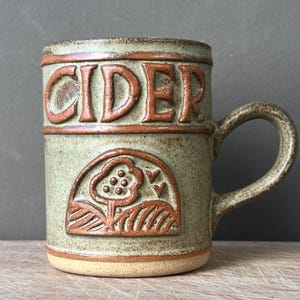 May include: A speckled light green ceramic mug with a brown handle and base. The word CIDER is embossed in brown letters near the top. Below the text, a brown embossed graphic depicts a tree with circular fruit and two birds in a field.