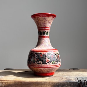 May include: A decorative ceramic vase with a speckled pink and beige body, accented with red bands and trim. The vase features a detailed design of mythical creatures in black, red, and green. The vase is displayed on a rustic wooden surface.