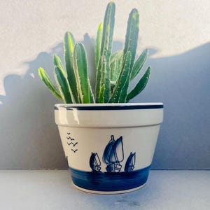 May include: A ceramic flower pot with a nautical design, featuring sailboats and birds in blue against a cream background. The pot holds a green cactus plant. The pot has a blue rim and base.