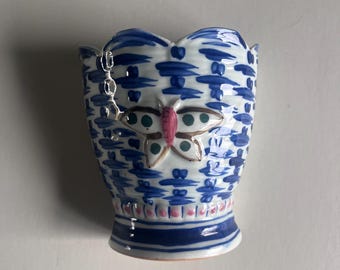 May include: A small, blue and white ceramic vase with a butterfly design. The vase has a scalloped rim and a flared base. The butterfly is pink, gold, and green. The vase is decorated with a blue and white pattern.