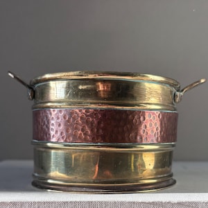 May include: A small, round metal container with a hammered copper band and brass accents. The container has two small handles and a slightly flared rim. The metal has a warm, aged patina, suggesting it is vintage or antique.