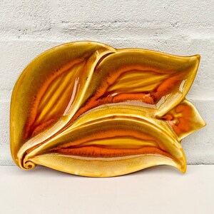 May include: A ceramic dish in the shape of a stylized flower with a yellow and orange glaze. The dish has a deep center and two overlapping petals.