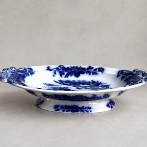 Op de afbeelding: Een ovale keramische serveerschaal met een witte basis en ingewikkelde blauwe bloemmotieven. De schaal heeft een verhoogde voet en decoratieve handvatten aan beide zijden, waarbij het blauwe bloemmotief zich uitstrekt over de binnen- en buitenkant.