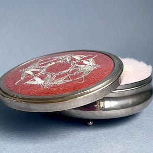 May include: An open, silver-toned powder compact with a red lid featuring an embroidered floral design. The compact contains a white powder puff. The compact is on a light blue surface.