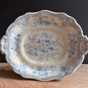 May include: An oval ceramic serving dish with a scalloped edge and two side handles. The off-white surface features a blue floral pattern with a central bouquet and a bird, complemented by a decorative border and a visible network of fine cracks.