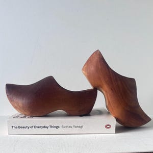 Puede incluir: Dos zuecos de madera, cada uno con un diseño curvo, se exhiben sobre un libro blanco titulado "The Beauty of Everyday Things". Los zuecos son de color marrón cálido, y el libro tiene una cubierta blanca con texto negro.