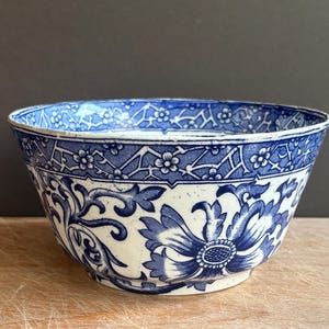 May include: A ceramic bowl featuring a white base with intricate blue floral patterns and a geometric border design around the rim. The exterior displays large stylised blossoms and scrolling foliage, creating a detailed contrast against the white surface.