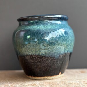 May include: A small, rounded ceramic pot featuring a glossy glaze that transitions from a deep, dark brown at the base to a vibrant, mottled teal and blue at the top. The rim is smooth and dark, with a visible light-coloured base edge.