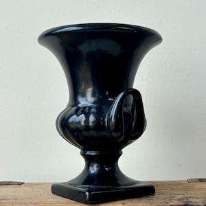 May include: A glossy black ceramic urn-style vase featuring a wide, flared rim, a rounded body with vertical ribbed detailing, and a curved handle on the side. The vessel stands on a square pedestal base with a smooth, reflective finish throughout.