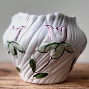 May include: A white ceramic vase with a textured, ribbed surface featuring embossed floral motifs. The flowers are painted with light green leaves and stems, accented by thin, dark pink outlines on the petals, creating a relief pattern.