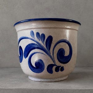 Può includere: Un vaso in ceramica con base bianco sporco maculata e bordo blu intenso. L'esterno presenta un motivo floreale blu dipinto a mano con viticci e foglie che avvolgono il corpo curvo del contenitore.