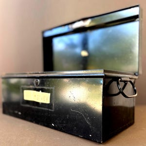 Könnte beinhalten: Ein rechteckiger Werkzeugkasten aus schwarzem Metall mit einem aufgeklappten Deckel. Der Kasten verfügt über einen kleinen schwarzen Griff an der Seite und ein gelbes rechteckiges Etikett auf der Vorderseite mit dem Text "Mr. Harvey" in schwarzer Schrift.