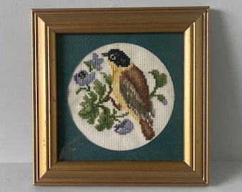 May include: A square gold-toned frame containing a circular cross-stitch embroidery of a bird with a black head, yellow chest, and brown wings perched on a branch with green leaves and small purple flowers against a dark green background.