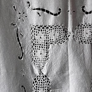 May include: A white textile featuring intricate cutwork embroidery and crochet lace detailing. The fabric displays a floral motif created through cutwork, complemented by a geometric crochet border that forms a corner pattern on the material.