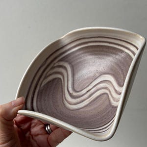 May include: A ceramic dish with a fan-like shape featuring a swirling pattern of muted purple and off-white tones. The bowl has a smooth, curved rim and a deep interior basin, showcasing a marbled aesthetic across the entire surface area.