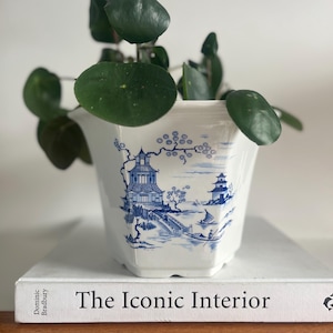 Puede incluir: Una maceta de cerámica blanca y azul con un diseño de pagoda, que contiene una planta verde con hojas redondas. La maceta está sobre un libro titulado "The Iconic Interior".