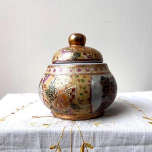 May include: A decorative ceramic jar with a lid. The jar is round with a gold base and lid. The jar is decorated with gold, purple, green, and white floral and geometric patterns. The jar is sitting on a white tablecloth with gold embroidery.
