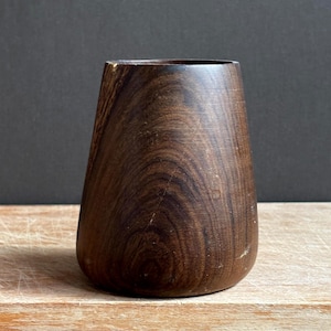 May include: A dark brown wooden cup with a tapered, cylindrical shape and a smooth, polished finish. The wood grain is visible throughout the exterior, displaying natural swirling patterns and deep brown tones in a simple, functional design.
