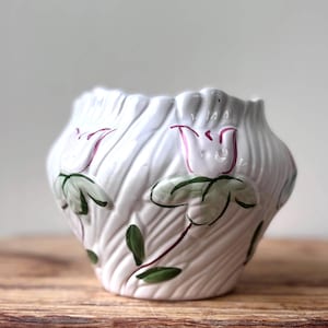 May include: White ceramic planter with a textured surface and floral design. The planter features pink and green painted flowers and leaves. The rim has a scalloped edge. The planter is sitting on a wooden surface.