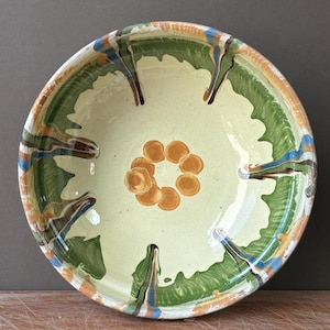 May include: A round ceramic bowl with a light cream-colored interior featuring a central cluster of seven orange circular spots. The rim and sides are decorated with green, blue, and brown drip-style patterns that extend toward the center.
