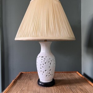 May include: A white ceramic table lamp with a cream pleated lampshade. The lamp base features a cut-out floral design and rests on a black base. The lamp is on a woven table.